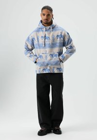 Blauwe capuchontrui met patroon van cactussen en paarden, gemaakt van zachte stof. Gecombineerd met zwarte broek met wijde pijpen en stevige zwarte schoenen.