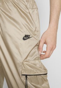 Bežnye najlončke cargo hlače, ki imajo stranski žep z črnim zadrgo, nastavljiv vrvico in črn Nike logotip na stegnu.
