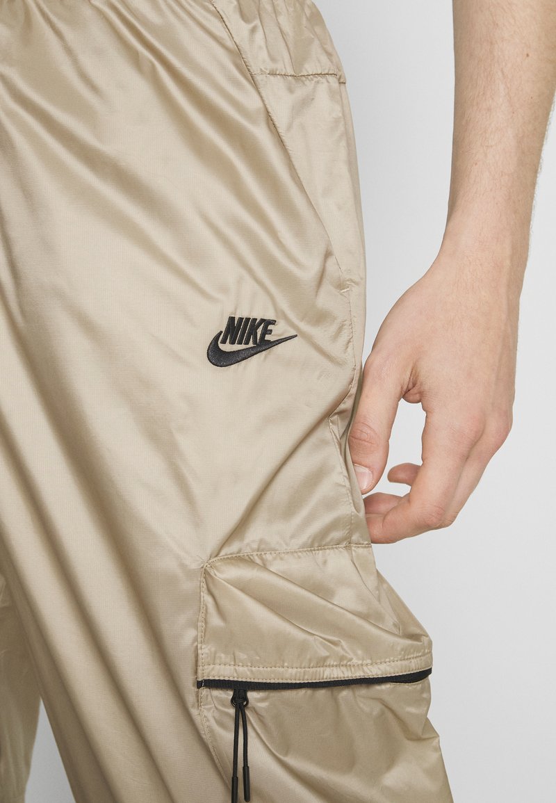 Pantalons cargo en nylon beige avec une poche latérale dotée d'une fermeture éclair noire, un cordon de serrage ajustable et un logo Nike noir sur la cuisse.