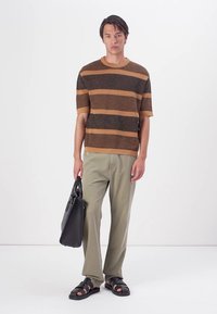 Bruine en tan gestreepte gebreide trui, lichtkhaki broek, zwarte tote bag en zwarte sandalen met bandjes. Model staat voor een effen achtergrond.
