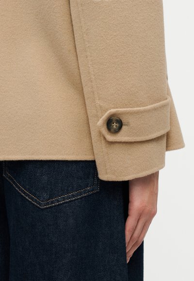 Μανίκι μπεζ παλτού από μαλλί με λεπτομέρεια κουμπωτού μανσετέ, απεικονιζόμενο με το χέρι να ακουμπά δίπλα στην τσέπη σκούρων μπλε τζιν παντελονιών.