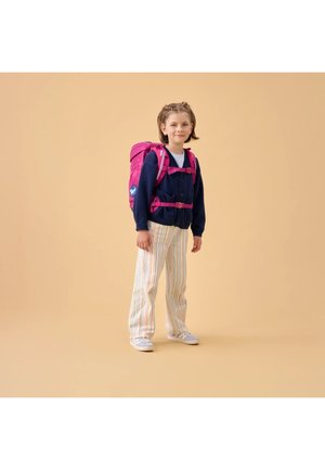 Child standing wearing a navy cardigan, striped pants, white sneakers, and a pink backpack with chest straps against a beige background.