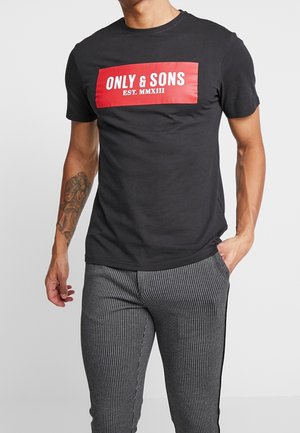 Homme portant un t-shirt noir avec un logo rectangulaire rouge indiquant "ONLY & SONS EST. MMXIII" et un pantalon gris à fines rayures, une main dans la poche.