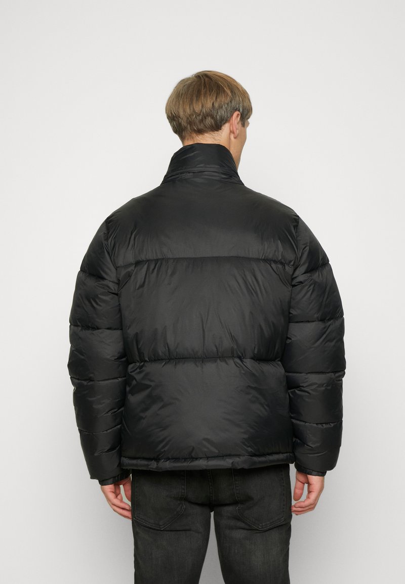 Doudoune noire avec un col haut, dotée de sections matelassées et d'une texture lisse. La vue arrière montre peu de détails de couture et des poignets élastiques.