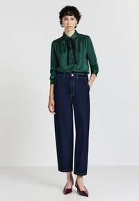 Blusa di satin verde con un papillon nero, abbinata a jeans in denim blu scuro a vita alta e scarpe con tacco borgogna.