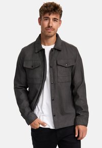 Chaqueta gris oscuro con un cuello, botones frontales y dos bolsillos en el pecho. La tela parece suave, complementando una camiseta blanca por debajo.