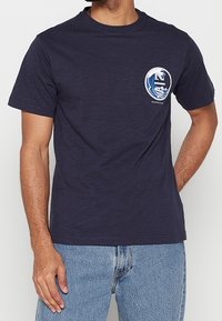 Granatowy bawełniany t-shirt z okrągłym dekoltem, z okrągłą grafiką fal i literą "E" w białym i niebieskim kolorze na lewej piersi.