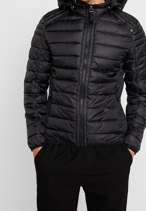 Man in een zwarte gewatteerde hoodie met ritszakken en zwarte broek, met handen in de zakken, tegen een effen achtergrond.