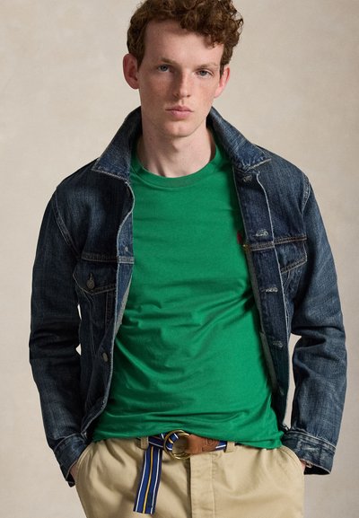 Ung mand med krøllet hår iført en grøn t-shirt, mørk cowboyjakke, beige bukser og et blåt stribet stofbælte med ringespænde.