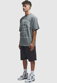 Szürke, oversized t-shirt fehér grafikus szöveggel és dizájnokkal, sötét rövidnadrággal és fekete akcentusokkal ellátott fehér sportcipővel párosítva.