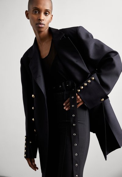 Black tailored blazer with gold button accents, featuring structured shoulders and a long, belted design. Paired with black fitted pants.