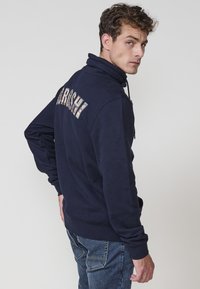 Sudadera azul marino con cuello alto, que presenta un texto gráfico multicolor en la parte posterior. Tejido suave, mangas largas y puños de canalé.