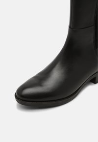 Botín negro de cuero hasta el tobillo con puntera puntiaguda, textura suave, costuras mínimas y un tacón cuadrado de contraste. Diseño elegante con elástico en los laterales.
