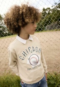 Sudadera beige con la palabra "Chicago" y un gráfico de béisbol en la parte frontal. Combinada con una camisa blanca con cuello debajo y pantalones cortos de mezclilla.
