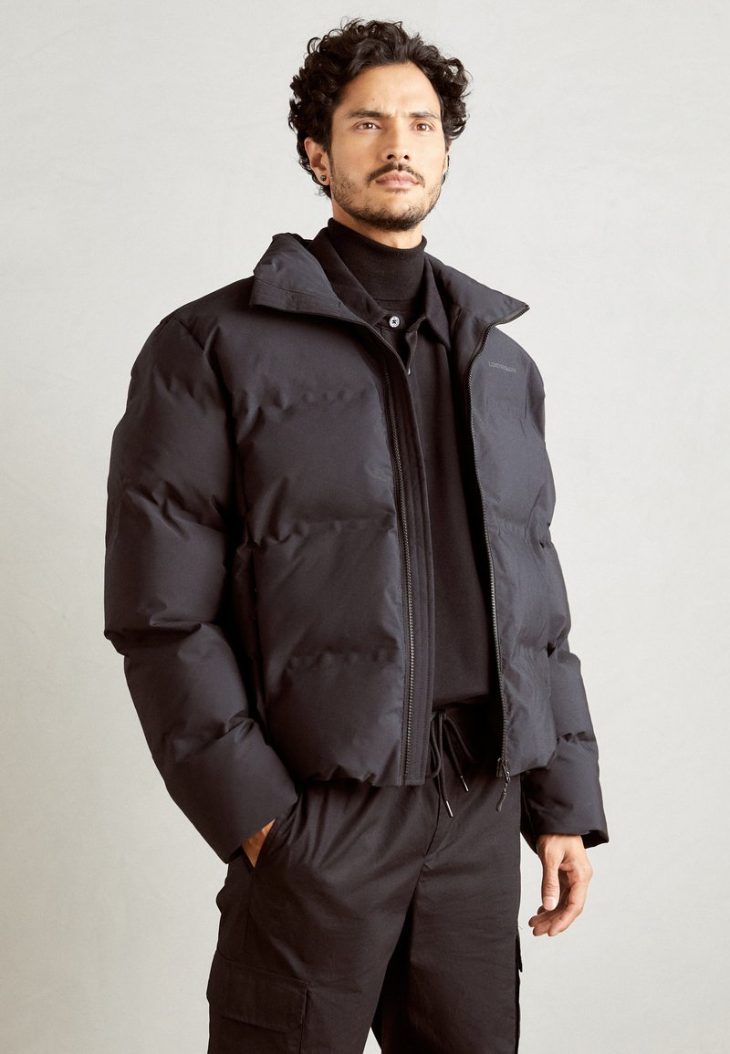 Doudoune noire au design court, avec fermeture éclair frontale, col haut et texture rembourrée. Portée sur un col roulé noir et un pantalon.