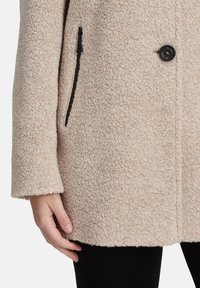 Beige fleece jas met een gestructureerd oppervlak, sluiting met één zwarte knop en zijzakken met ritssluiting. Lange mouwen met een rechte zoom.