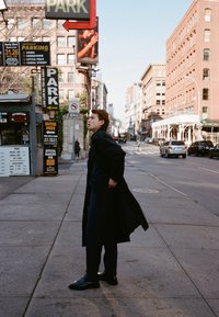 Muž v dlouhém černém kabátu kráčí po chodníku poblíž značek parkoviště a městské ulice s auty a budovami za denního světla.