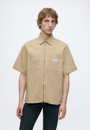 Jeune homme aux cheveux bouclés portant une chemise beige à manches courtes avec fermeture éclair et poches avant, ainsi qu'un pantalon foncé, sur un fond uni.