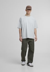 Világoskék, oversized póló, olivazöld nadrág zsebekkel és barna sapka felirattal. Fehér sportcipők szürke részletekkel és fekete talppal.