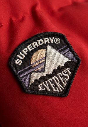 Patch ricamato in bianco e nero con montagne, un sole e il testo "SUPERDRY", su uno sfondo di tessuto rosso. Forma esagonale con design dettagliato.