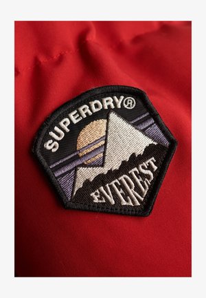 Patch ricamato in bianco e nero con montagne, un sole e il testo "SUPERDRY", su uno sfondo di tessuto rosso. Forma esagonale con design dettagliato.