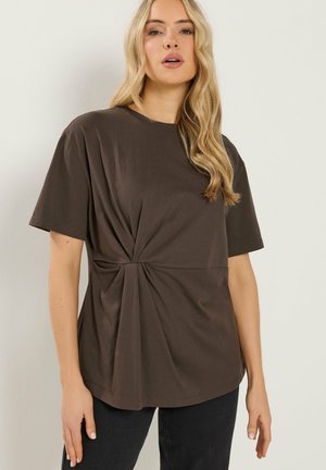 Lys blond kvinde iført løs, mørkebrun kortærmet top med samlet detalje i siden, kombineret med sorte bukser, stående mod en lys væg.