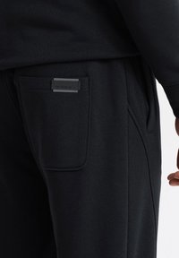 Pantalones de chándal negros con un acabado texturizado, que cuentan con un bolsillo lateral con una etiqueta negra y una cinturilla elástica para mayor comodidad.