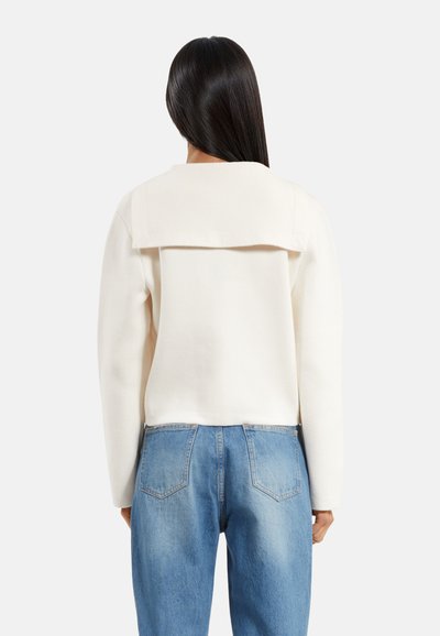 Kvinde med langt mørkt hår iført en hvid langærmet bluse med bred krave og blå jeans, stående med ryggen til mod en ensfarvet baggrund.