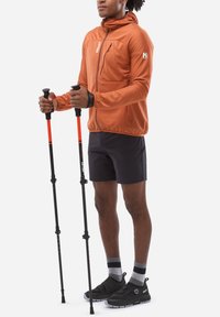 Veste zippée orange avec finition texturée, shorts noirs et chaussures noires. Deux bâtons de trekking avec des accents orange tenus à la main.
