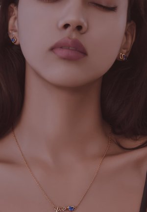 Collier en or avec "amour" en cursive et un pendentif en forme de cœur bleu. Boucles d'oreilles en or ornées de petites pierres bleues. Fond de teint.