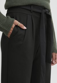 Pantalon noir taille haute avec plis sur le devant et ceinture nouée, fabriqué en tissu lisse, associé à un pull en tricot vert.