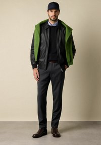 Man in zwarte leren jas, groene trui over zijn schouders, donkere broek, zwarte pet en bruine laarzen die tegen een neutrale achtergrond staat.