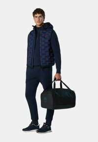 Marineblauer Puffer-Weste über einem passenden Trainingsanzug, beide aus weichem Stoff gefertigt. Schwarze Sporttasche mit Logo. Sneakers mit dunkelblauen und braunen Akzenten.