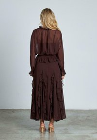 Top lungo marrone trasparente con maniche a sbuffo e collo arricciato, abbinato a una gonna a lunghezza intera con strati di rouches e un design fluido.