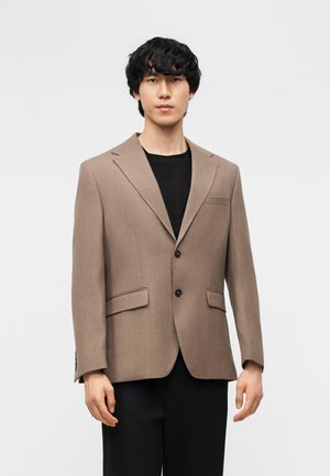 Man die een beige blazer draagt over een zwart overhemd en zwarte broek, staand tegen een effen lichtgrijze achtergrond.