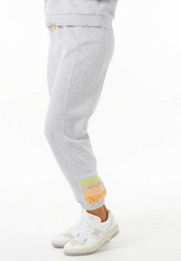 Persona vestida con pantalones de chándal gris claro con detalles de puños multicolor y zapatillas blancas, de pie contra un fondo blanco.