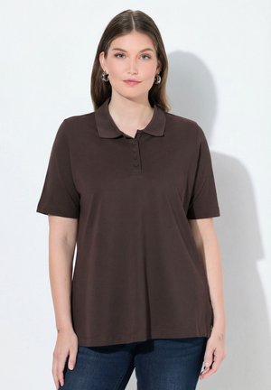 Žena v hnědém polo tričku s krátkým rukávem a modrých džínách stojí před jednoduchým světlým pozadím.