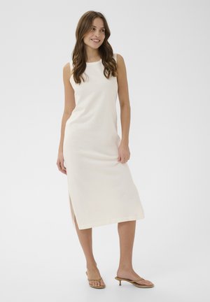 Donna sorridente con lunghi capelli mossi che indossa un abito midi bianco senza maniche e sandali beige con tacco, in piedi davanti a uno sfondo semplice.