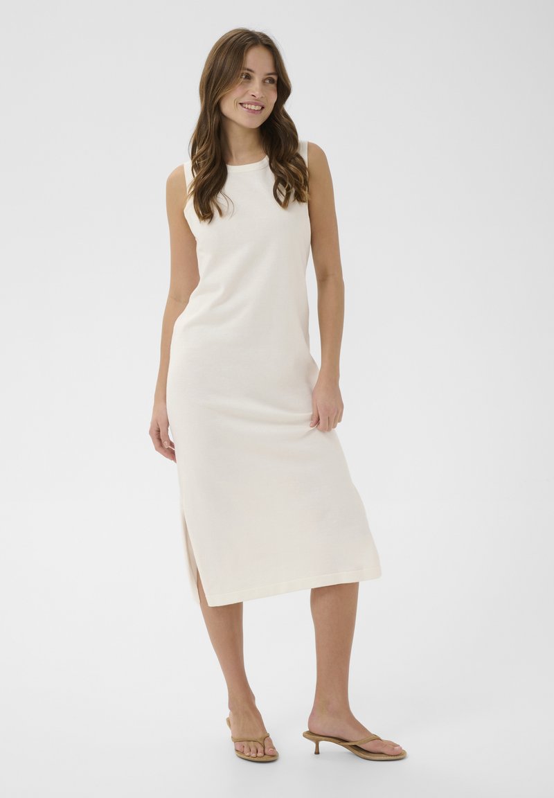 Femme souriante aux longs cheveux ondulés, portant une robe midi blanche sans manches et des sandales à talons beiges, debout devant un fond uni.