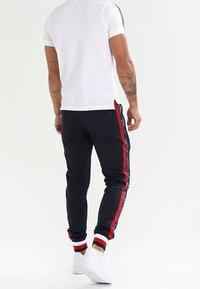 Witte polo met korte mouwen, gecombineerd met marineblauwe joggingbroek met rode en witte zijstrepen. Het materiaal lijkt glad en aansluitend.