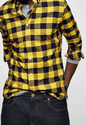 Homme portant une chemise à carreaux jaune et noire avec les manches retroussées par-dessus un t-shirt henley gris et un jean bleu foncé.