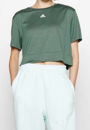 Osoba ubrana w zielony krótki t-shirt Adidas i białe spodnie z elastycznym pasem, z rękami w kieszeniach na jednolitym tle.