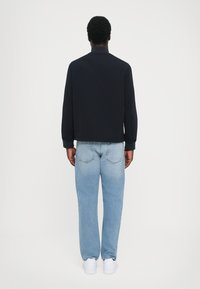 Sort jakke med høj krave, tætsiddende manchetter, kombineret med lyseblå afslappede jeans og hvide sneakers. Bagfra, minimalistisk design.