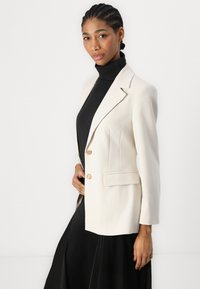 Samanvärinen blazer med två guldknappar, slagkrage och två sidfickor. Stylad över en svart polotröja, med raka linjer och skräddarsydd passform.