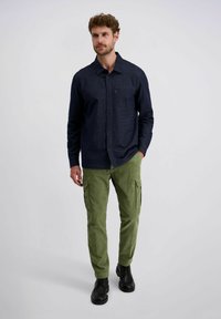 Man in een donkerblauwe knopshirt, olijfgroene cargo broek en zwarte schoenen, staand met één hand in zijn zak tegen een effen achtergrond.