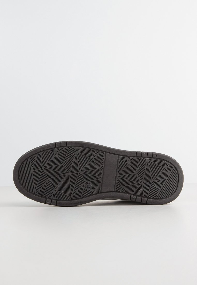 Suola di una scarpa nera con motivi di battistrada geometrici che includono triangoli e linee, esposta su una superficie bianca contro uno sfondo neutro.