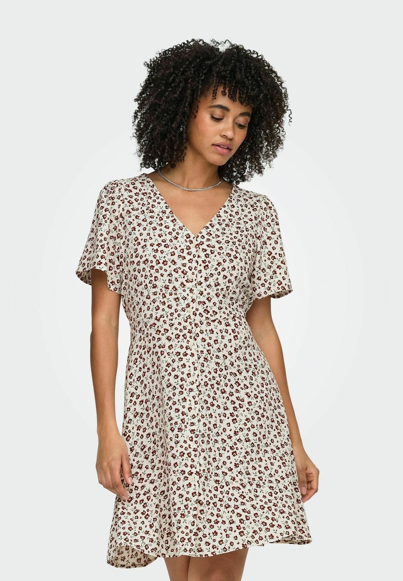 Femme aux cheveux bouclés portant une robe blanche à manches courtes, col en V, longueur genou, avec un petit motif floral rouge.