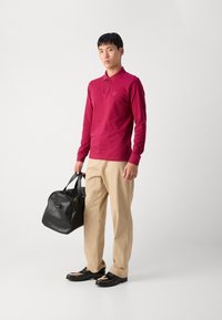 Bordeaux lange mouwen polo met knoopkraag, beige broek, glanzende zwarte schoenen en een gestructureerde zwarte leren tas.