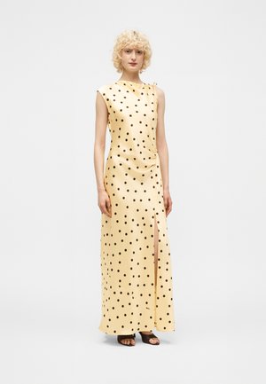 Γυναίκα με ξανθά σγουρά μαλλιά που φοράει κίτρινο σατέν φόρεμα χωρίς μανίκια με μαύρα πουά και σχίσιμο μπροστά, στέκεται μπροστά σε απλό φόντο.