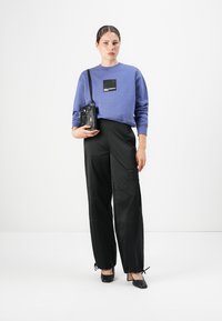 Woman standing wearing a blue sweatshirt, black wide-leg pants with drawstring hems, black heels, and holding a small black shoulder bag.